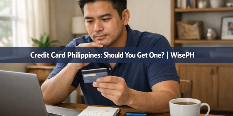 A young Filipino man thoughtfully holding a credit card at his home desk, weighing the decision of whether to get one.